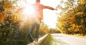 Henkilö kävelee ja tasapainoilee kädet levällään tien vieresä olevan matalan kaiteen päällä. Syksyistä metsää ja aurinko.