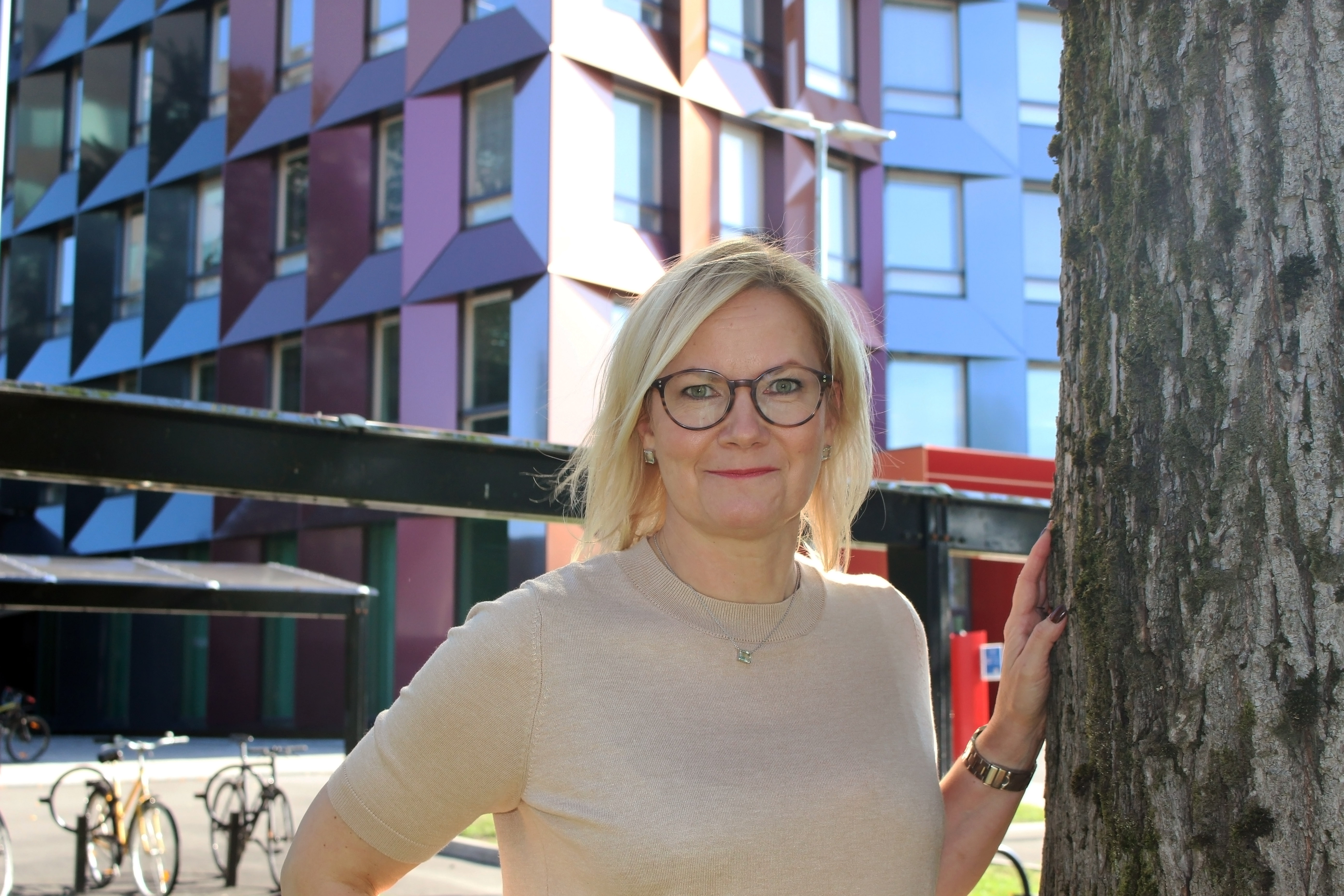 Katja katsoo aurinkoisessa säässä kameraan ja pitää kättä vasemmalla puolellaan olevan puun runkoa vasten.