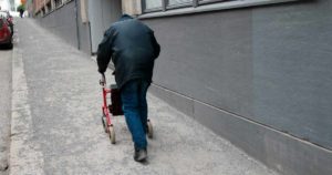 Henkilö kävelee harmaata katua ylöspäin rollaattorin avulla, maassa asfaltti ja hiekkaa