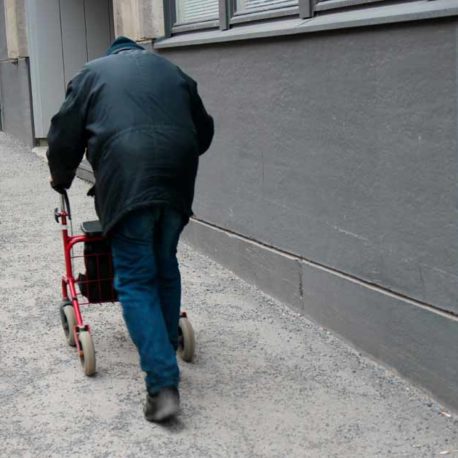Henkilö kävelee harmaata katua ylöspäin rollaattorin avulla, maassa asfaltti ja hiekkaa