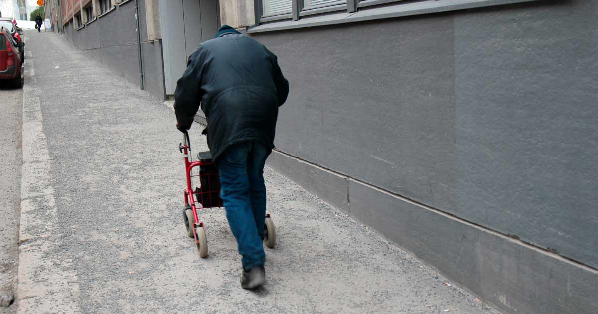 Henkilö kävelee harmaata katua ylöspäin rollaattorin avulla, maassa asfaltti ja hiekkaa