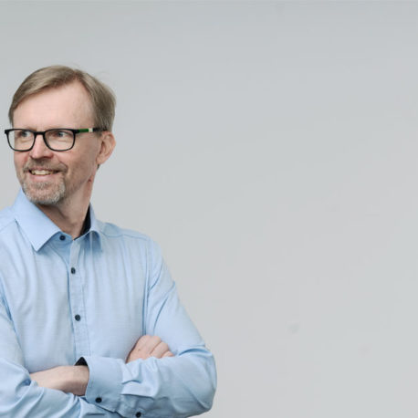 Mikko katsoo kuvassa vasemmalle, kädet puuskassa, harmaa tausta ja Mikolla sininen kauluspaita.