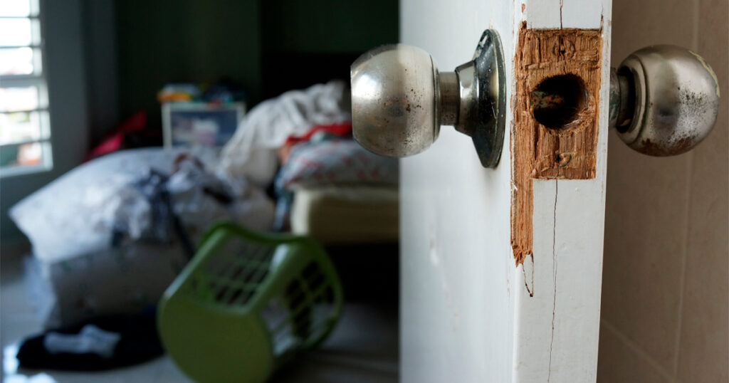 Kuvassa ovi, jossa murron jälkiä. Taustalla huone, jossa tavarat hujan hajan.